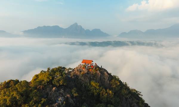 Eine grüne Bergkuppe ragt in die Wolken, auf ihrer Spitze ein rotes Wellblechdach, daneben eine Gruppe Menschen, die in die Ferne schaut.
