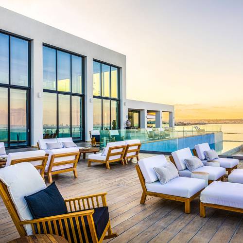Terrasse einer modernen Hotelanlage mit Holzliegen und wei&szlig;en Auflagen, Blick auf das Meer und den Sonnenuntergang, gro&szlig;e Fensterfront.