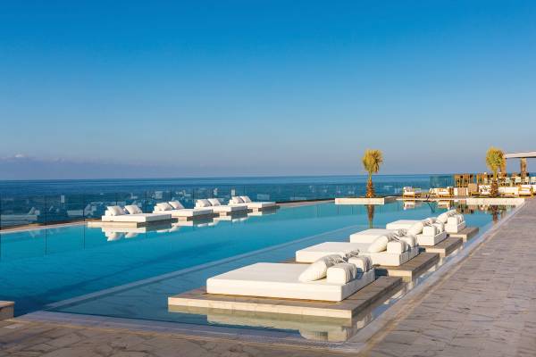 Hotelanlage mit Infinity-Pool, wei&szlig;en Liegen und Palmen am Rand. Der Pool spiegelt den blauen Himmel und das Meer wider.