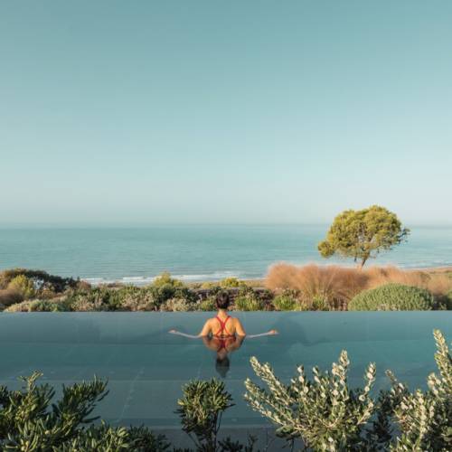 Person im roten Badeanzug entspannt im Infinity-Pool mit Blick auf das Meer und K&uuml;stenlandschaft