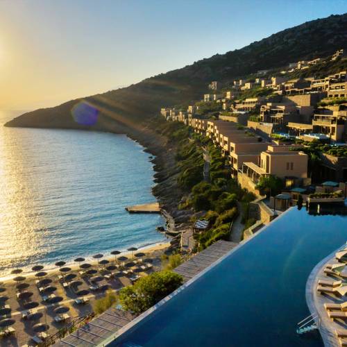 Luftaufnahme einer Hotelanlage am Meer mit mehreren Geb&auml;uden auf einem Hang, einem Infinity-Pool und Liegen am Strand. Sonnenuntergang im Hintergrund.