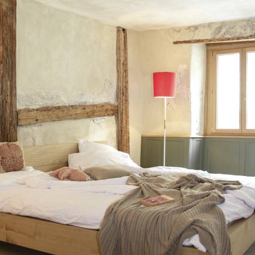 Ein helles Hotelzimmer mit einem gro&szlig;en Holzbett, wei&szlig;en Bettlaken und einer grauen Decke. Eine rote Stehlampe steht neben dem Fenster. Im Hintergrund ist eine Person in Bewegung.
