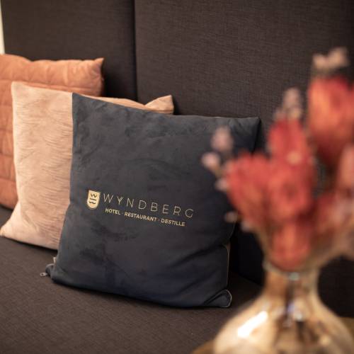 Dunkelblaues Kissen mit dem Schriftzug 'WYNDBERG HOTEL, RESTAURANT, DESTILLERIE' auf einer Couch mit zwei hellen Kissen und einer Vase mit Blumen.