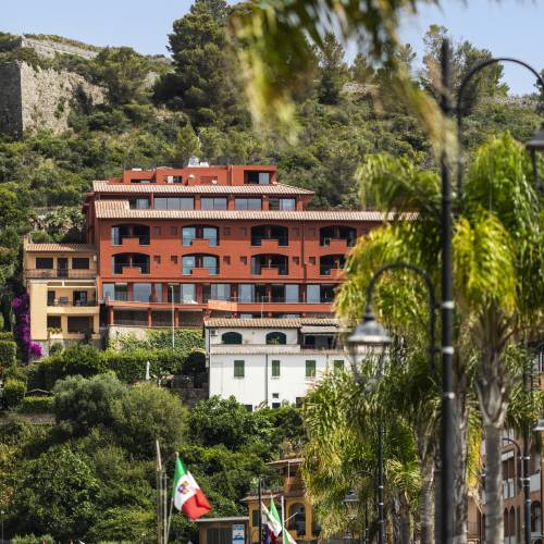 Mehrst&ouml;ckiges Hotelgeb&auml;ude in Rot und Beige an einem Hang mit Blick auf eine Promenade mit Palmen und Flaggen, umgeben von gr&uuml;ner Vegetation
