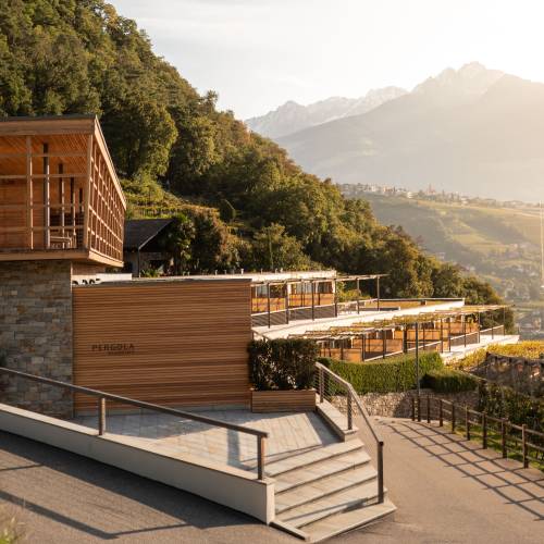 Moderne Hotelanlage mit Holz- und Steinfassade, umgeben von B&auml;umen und Weinreben. Im Hintergrund Berge und Sonnenuntergang.