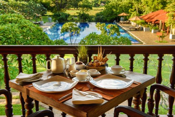Fr&uuml;hst&uuml;ckstisch mit Tassen, Teekanne und Geb&auml;ck auf der Terrasse des Liyawela Hotels mit Blick auf den Pool und Garten