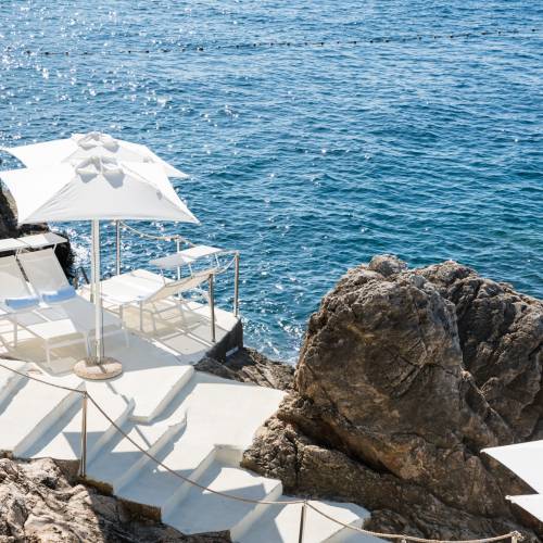 Hotelterrasse mit wei&szlig;en Liegest&uuml;hlen und Sonnenschirm direkt am Felsen &uuml;ber dem Meer