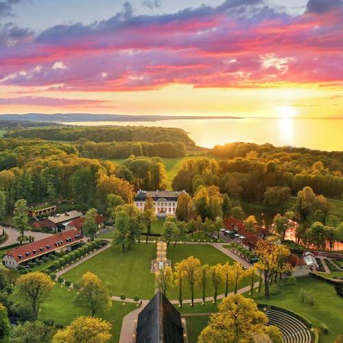 Luftaufnahme einer Hotelanlage mit mehreren Geb&auml;uden, gepflegten G&auml;rten und B&auml;umen, umgeben von Wasser und einem Sonnenuntergang im Hintergrund.