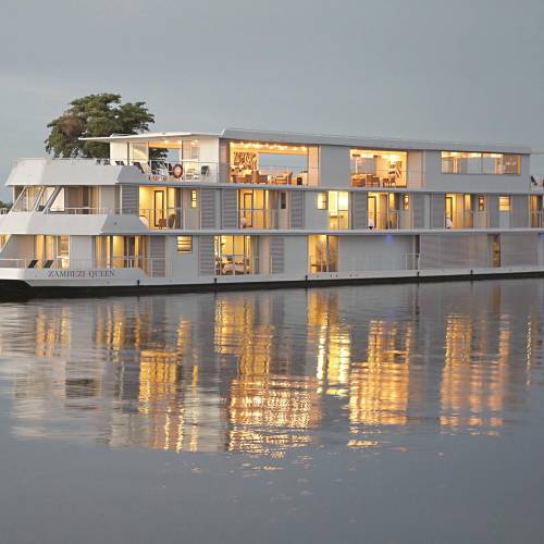 Das Hotelboot 'Zambezi Queen' schwimmt auf ruhigem Wasser, mit mehreren Fenstern, die warmes Licht ausstrahlen. Umgeben von gr&uuml;ner Vegetation.