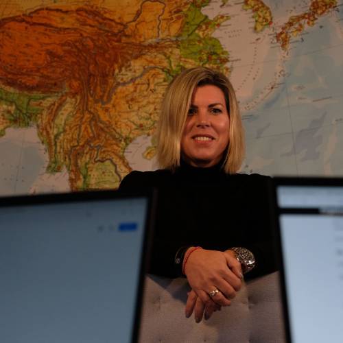 Christine Elsner steht schwarz gekleidet vor einer gro&szlig;en Landkarte und l&auml;cheln freundlich in die Kamera. Im Vordergrund sind links und rechts von ihr Computerbildschirme zu sehen.