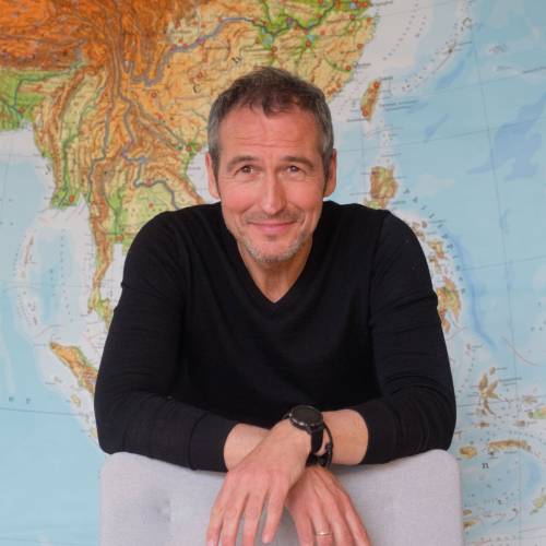Henrik He&uuml;veldop lehnt mit verschr&auml;nkten H&auml;nden &uuml;ber der R&uuml;ckenlehne eines hellen Stuhls, er schaut freundlich in die Kamera. Im Hintergrund ist eine gro&szlig;en Landkarte zu sehen.