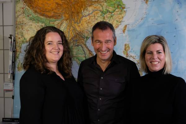 Sharlyn Tripp, Henrik He&uuml;veldop und Christine Elsner stehen schwarz gekleidet vor einer gro&szlig;en Landkarte und l&auml;cheln freundlich in die Kamera.